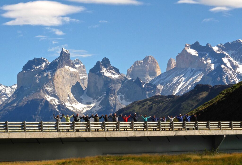 A group of hikers standing on a bridge in Torres del Paine National Park, with the Cuernos and other mountains behind them