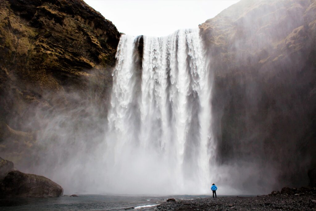 Person standing on rocky land in front of a mist created by a large waterfall crashing in a body of water