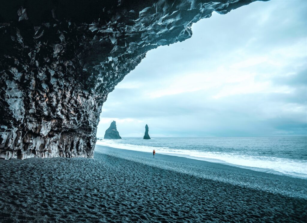 Person walking along the shore of a black-sanded beach near a rocky cave in Iceland