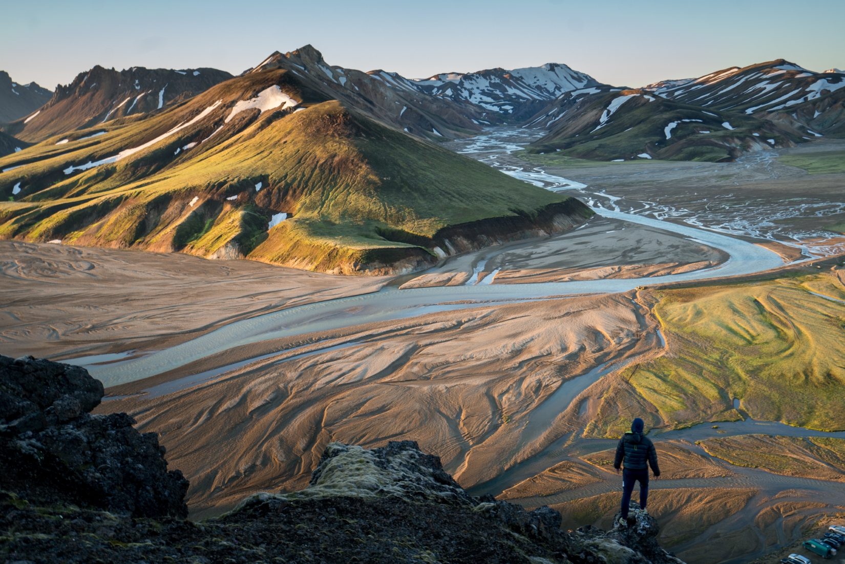 Man standing on a cliff looking at a valley of snow-dusted mountains and paths made by water.